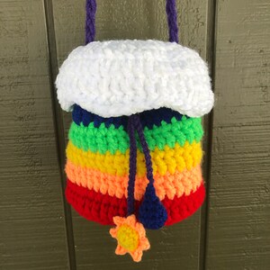 May include: A rainbow-colored crocheted drawstring bag with a white crocheted cloud top. The bag has a purple cord and a yellow crocheted sun charm.
