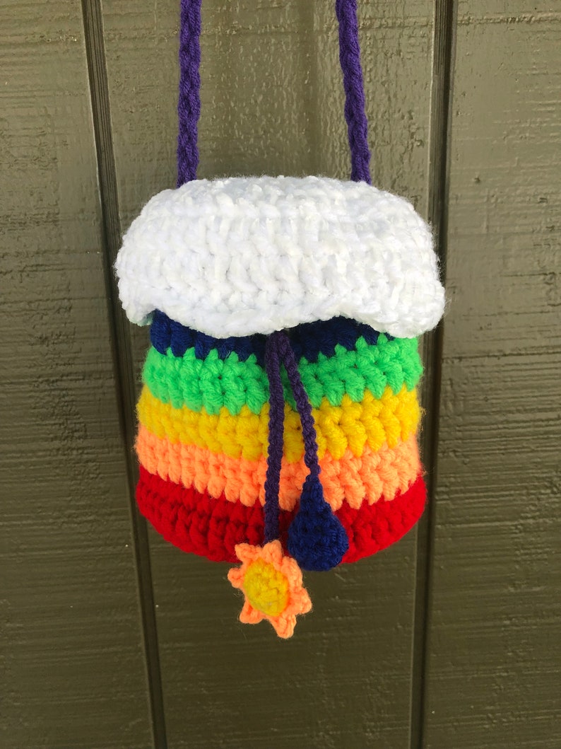 May include: A rainbow-colored crocheted drawstring bag with a white crocheted cloud top. The bag has a purple cord and a yellow crocheted sun charm.