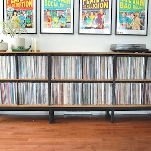 May include: A wooden record storage cabinet with black metal legs, filled with vinyl records. The cabinet has multiple shelves and is topped with a record player. Four framed posters hang above the cabinet.