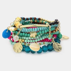 Puede incluir: Un conjunto de pulseras de cuentas coloridas. Las pulseras presentan cuentas en tonos azules, verdes, rosas y dorados. Algunas pulseras tienen dijes y colgantes dorados, incluyendo una mariposa y discos redondos. Las pulseras están apiladas juntas.