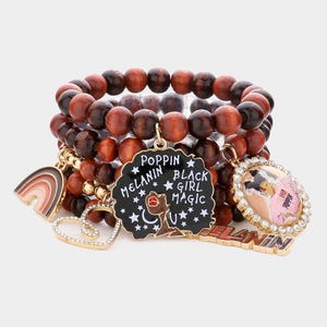 Puede incluir: Un conjunto de cuatro pulseras de cuentas de madera marrón con dijes de oro. Los dijes incluyen un corazón, un arcoíris, un círculo blanco y negro con el texto "Poppin Melanin Black Girl Magic", y un dije dorado con el texto "Melanin" y un retrato de una mujer.