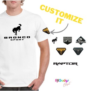 May include: White t-shirt with a black Bronco Sport logo featuring a rearing horse. The shirt has a customizable patch on the sleeve.  The image shows five different patch options: a black horse, a gray mountain range, a gray wave, a yellow diamond with a black horse, and a yellow diamond with a black horse and the words "Bronco Sport". The text "CUSTOMIZE IT" and "RAPTOR" are also visible.