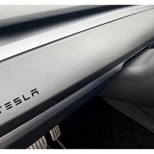 May include: Close-up of a black dashboard in a car with the word "TESLA" in black lettering.