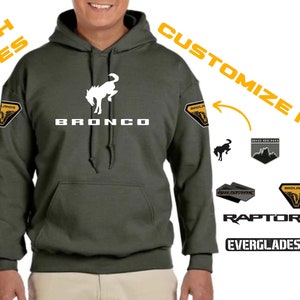 May include: A green hooded sweatshirt with a white Bronco logo on the front. The sleeves have patches that can be customized with different designs, including "Wildtrak", "Raptor", "Everglades", "Black Diamond", "Outer Banks", and "Sahara Side Steps".