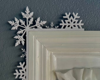 May include: White snowflake decorations are placed on the corner of a white window frame. The window has white sheer curtains.