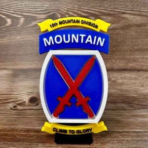 May include: A 3D-printed emblem of the 10th Mountain Division. The emblem features a blue and white oval with two crossed red swords. The words "MOUNTAIN" and "CLIMB TO GLORY" are displayed.