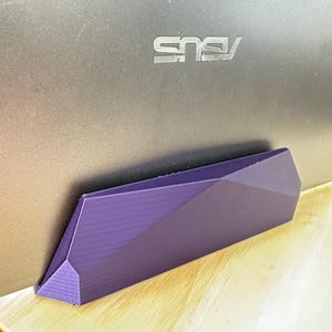 May include: A purple, 3D-printed laptop stand with a geometric, faceted design. The stand features a long, angular shape that supports a dark gray ASUS laptop, showcasing a textured surface finish consistent with additive manufacturing processes.