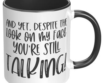 May include: White ceramic mug with a black interior and handle. The mug features the text "AND YET, DESPITE THE LOOK ON MY FACE YOU'RE STILL TALKING!" in black lettering. A humorous gift idea.