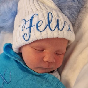 May include: A white knitted baby hat with a blue pom-pom and the name "Felix" embroidered in blue. The hat is on a baby's head.