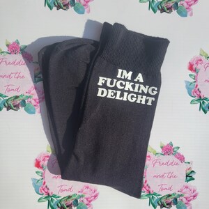 May include: Black socks with the text "IM A FUCKING DELIGHT" in white. The socks are folded and displayed on a floral background with the text "Freddie and the Toad".