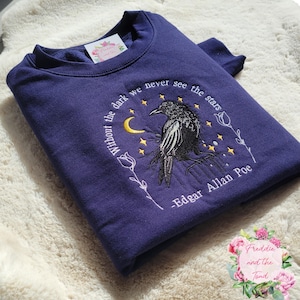May include: A navy blue sweatshirt with an embroidered design featuring a crow, crescent moon, stars, and floral accents. The text reads "Without the dark we never see the stars - Edgar Allan Poe."