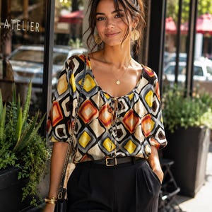 May include: A short-sleeved blouse featuring a geometric diamond pattern in shades of yellow, orange, brown, and grey on a cream background, paired with black trousers and a black belt with a gold buckle, accessorized with gold earrings and necklaces.