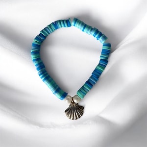 Könnte beinhalten: Ein blau-türkisfarbenes Perlenarmband mit einem goldenen Muschelanhänger und zwei weißen Perlen. Das Armband besteht aus flachen, runden Perlen in Blau- und Türkistönen. Der Muschelanhänger sorgt für einen maritimen Touch.