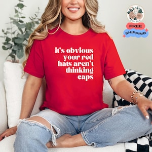 May include: A red t-shirt with the text "it's obvious your red hats aren't thinking caps" in white. The shirt is worn with blue jeans. The image also includes a bracelet and a tattoo.