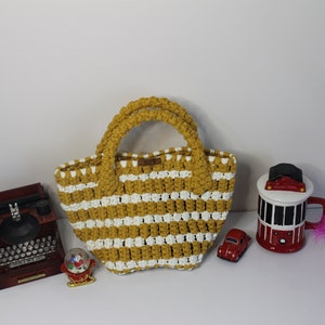 Könnte beinhalten: Eine gelbe und weiße gestreifte gehäkelte Tragetasche mit zwei Henkeln. Die Tasche liegt auf einer weißen Oberfläche mit einer alten Schreibmaschine, einer Schneekugel, einem roten Spielzeugauto und einer rot-schwarzen Tasse mit einer weißen Feder.