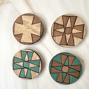 May include: Four circular wooden coasters featuring geometric inlay patterns. Two coasters display natural wood tones in light and dark brown, while the other two incorporate teal-colored wood segments arranged in symmetrical, angular designs.