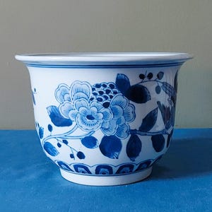 Pode incluir: Um vaso de cerâmica branca com um padrão floral azul pintado à mão, apresentando flores, folhas e bagas detalhadas. O recipiente em forma de taça tem uma borda larga e alargada e um design decorativo festonado na base do exterior.