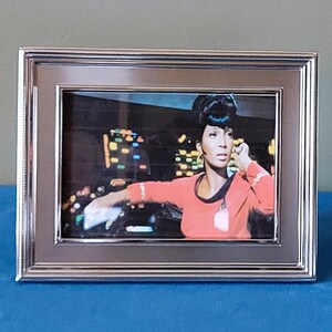 May include: A rectangular silver-toned metal picture frame containing a photograph of a person with dark hair wearing a coral-colored long-sleeved top. The person is positioned in front of a dark console with illuminated buttons and displays.