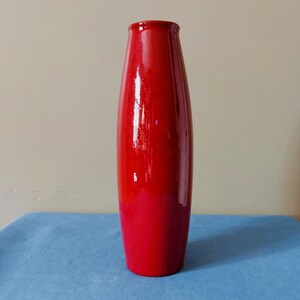 Peut inclure: Un grand vase cylindrique en céramique avec une finition lisse et brillante d'un rouge vif et uni. Le vase présente un col légèrement effilé et un bord arrondi, se tenant droit avec une forme cohérente et uniforme de la base à l'ouverture.