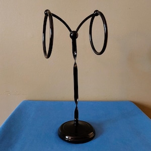 May include: A black metal towel holder with two circular rings. The holder has a twisted stem and a round base. The towel holder is on a blue surface.