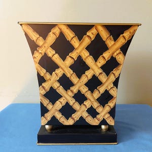 Peut inclure: Une corbeille à papier rectangulaire et évasée avec un fond noir orné d'un motif en treillis de bambou peint en beige et marron. Le récipient possède un bord doré, repose sur une base rectangulaire noire et quatre petits pieds dorés.