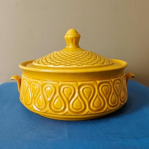 May include: A yellow ceramic casserole dish with a lid. The dish has a textured lid and a repeating teardrop pattern around the base. The dish has two small handles on either side.