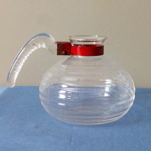 May include: A clear glass pitcher with a rounded body and a clear handle. The pitcher has horizontal ridges and a red band around the top. The handle is clear and attached with a rusty metal bracket.