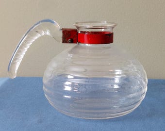 Op de afbeelding: Een heldere glazen kan met een afgeronde body en een heldere handgreep. De kan heeft horizontale ribbels en een rode band rond de bovenkant. De handgreep is helder en bevestigd met een roestige metalen beugel.