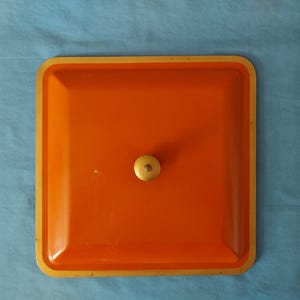 May include: An orange square tray with a gold rim and a small, round, light-colored knob in the center. The tray is set against a blue background.