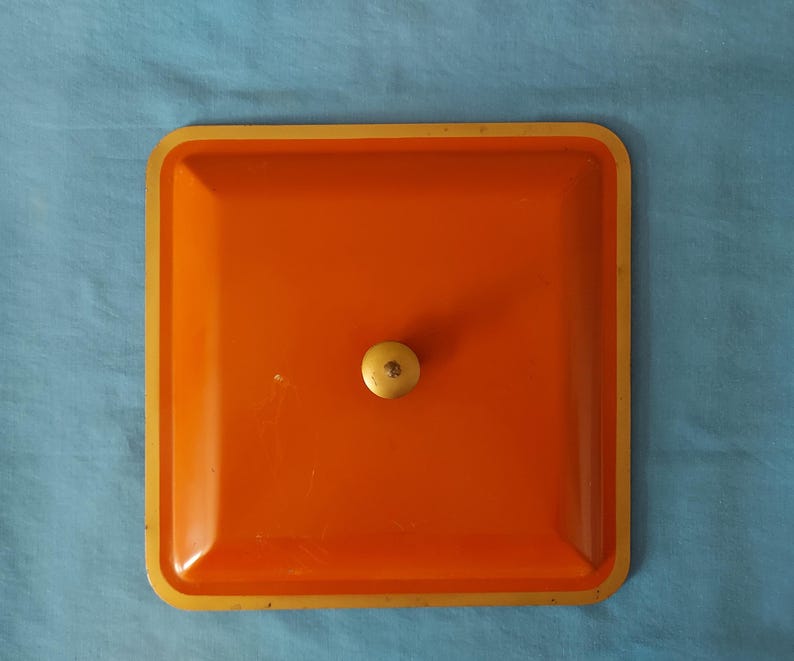 May include: An orange square tray with a gold rim and a small, round, light-colored knob in the center. The tray is set against a blue background.