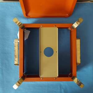 May include: An orange and gold metal camping stove with a matching lid. The stove has a square frame with four folding legs and a central beige panel with a circular cutout. The lid is open, revealing the interior.