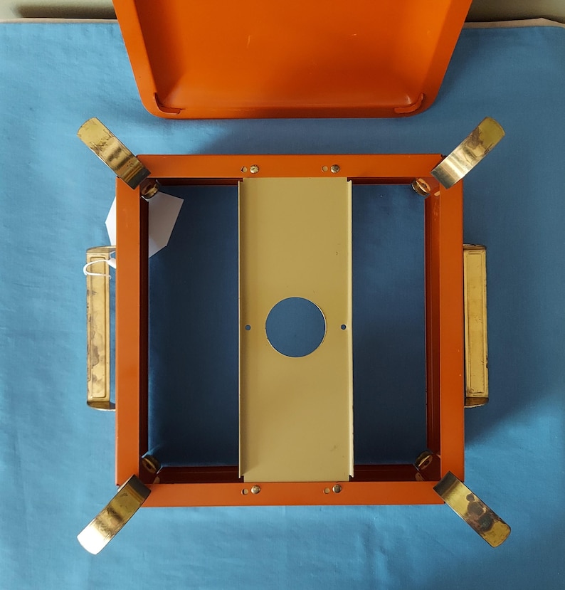 May include: An orange and gold metal camping stove with a matching lid. The stove has a square frame with four folding legs and a central beige panel with a circular cutout. The lid is open, revealing the interior.