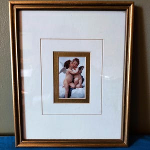 May include: A framed print featuring two cherubs embracing. The print is set within a white mat and a gold-colored frame. The cherubs are depicted in a classical style, with wings and a soft, warm color palette.