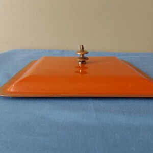 May include: An orange square lid with a bronze-colored rim and a small, decorative handle. The lid is set on a blue surface. The handle is a series of stacked, rounded shapes.