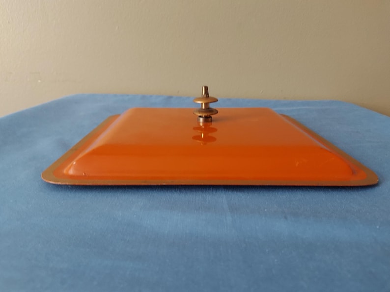 May include: An orange square lid with a bronze-colored rim and a small, decorative handle. The lid is set on a blue surface. The handle is a series of stacked, rounded shapes.