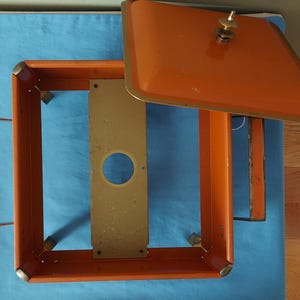 May include: An orange metal box with a matching lid, featuring a brass handle and trim. The box has a rectangular frame with a gold-colored metal plate inside. The lid is slightly ajar, revealing the interior.