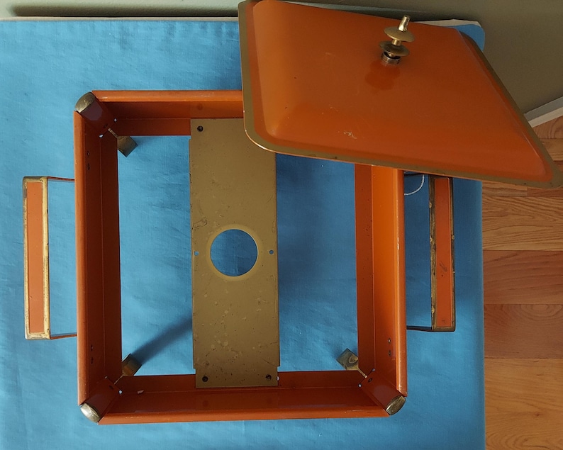 May include: An orange metal box with a matching lid, featuring a brass handle and trim. The box has a rectangular frame with a gold-colored metal plate inside. The lid is slightly ajar, revealing the interior.