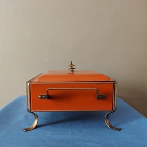 May include: An orange rectangular box with a handle and brass-colored trim and feet. The lid has a decorative finial. The box is sitting on a blue surface.