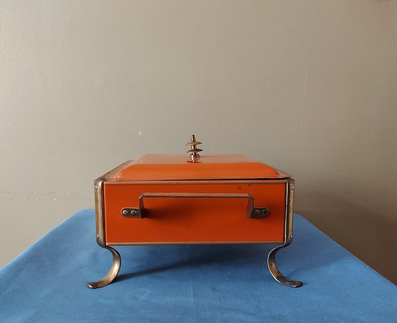 May include: An orange rectangular box with a handle and brass-colored trim and feet. The lid has a decorative finial. The box is sitting on a blue surface.