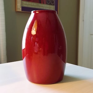 Peut inclure: Un vase en céramique rouge profond et brillant avec une silhouette lisse, arrondie et en forme de tonneau. La surface présente une finition réfléchissante avec un reflet lumineux marqué près du sommet, soulignant la structure courbe.