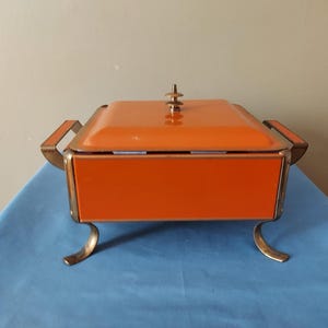 May include: An orange metal serving dish with a lid, handles, and decorative feet. The dish has a rectangular shape and a gold-colored trim. The lid is topped with a small, decorative finial. The dish is sitting on a blue surface.