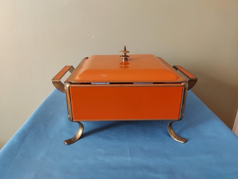 May include: An orange metal serving dish with a lid, handles, and decorative feet. The dish has a rectangular shape and a gold-colored trim. The lid is topped with a small, decorative finial. The dish is sitting on a blue surface.