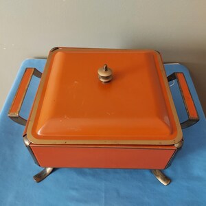 May include: An orange, square-shaped vintage serving dish with a lid and brass-colored accents. The dish has a handle on the lid and two side handles. It sits on four brass-colored feet. The dish is likely used for serving food.