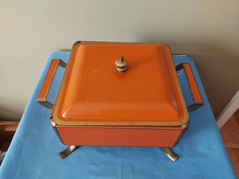 May include: An orange, square-shaped vintage serving dish with a lid and brass-colored accents. The dish has a handle on the lid and two side handles. It sits on four brass-colored feet. The dish is likely used for serving food.