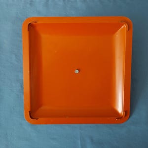 May include: An orange square tray with rounded corners and a small metal fastener in the center. The tray has a slightly recessed surface and a smooth, glossy finish. The background is a solid blue color.