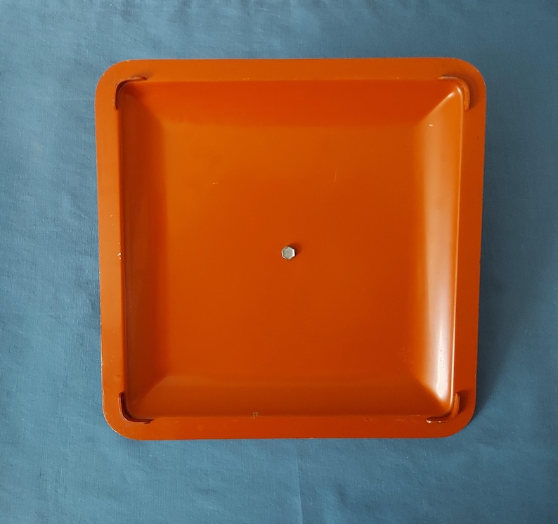 May include: An orange square tray with rounded corners and a small metal fastener in the center. The tray has a slightly recessed surface and a smooth, glossy finish. The background is a solid blue color.