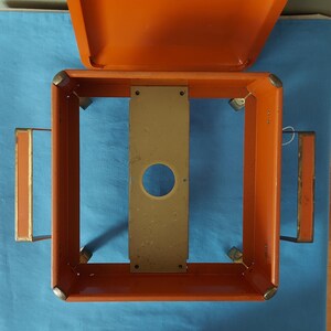 May include: An orange metal box with a hinged lid, open to reveal a rectangular frame. The frame has a central, tan-colored plate with a circular hole. Two orange handles with tan accents are on the sides.
