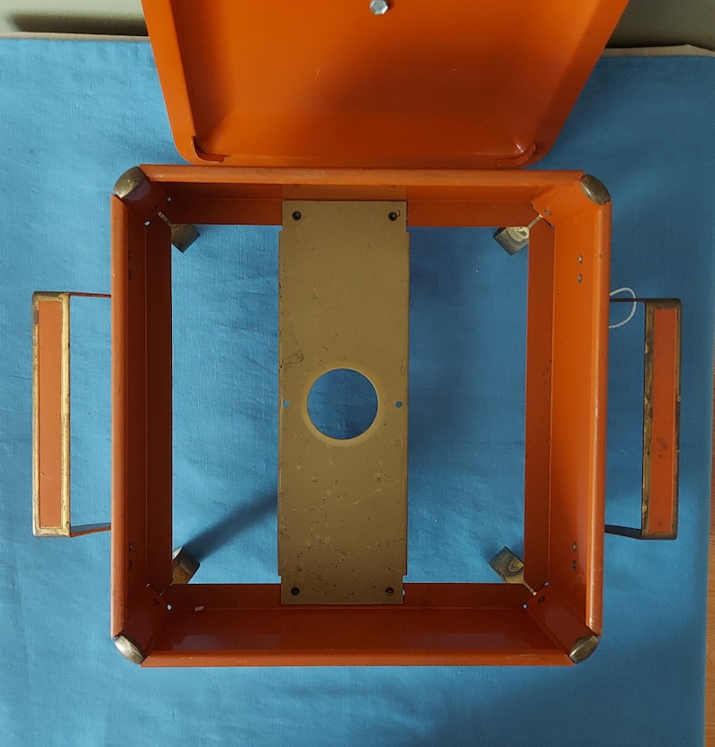 May include: An orange metal box with a hinged lid, open to reveal a rectangular frame. The frame has a central, tan-colored plate with a circular hole. Two orange handles with tan accents are on the sides.