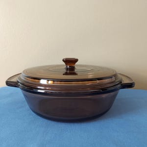 May include: A brown glass casserole dish with a lid. The dish is round and has two handles on the sides. The lid has a small knob on top.
