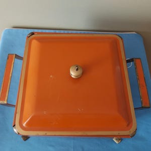 May include: An orange metal serving dish with a square shape and a lid. The dish has a gold trim and handles, and a small, off-white knob on the lid. The dish is sitting on a blue surface.